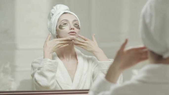 穿着浴袍和毛巾的年轻女子，看着镜子里的自己，用手将保湿霜涂在脖子和脸上。