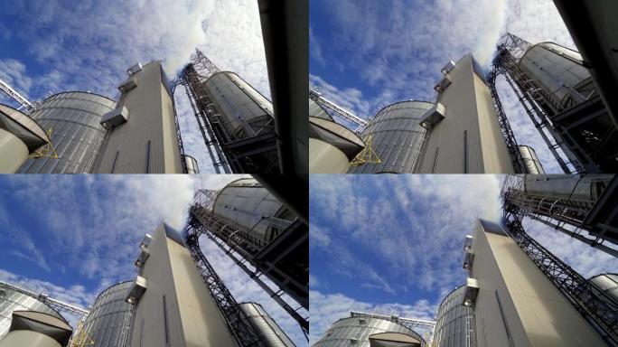 蓝天下的谷物升降机。工业建筑释放出浓浓的白烟。农业综合企业的现代综合体。从下面看。
