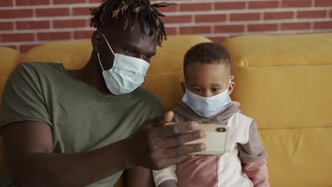 父子俩戴着医用口罩坐在沙发上，在智能手机上观看视频