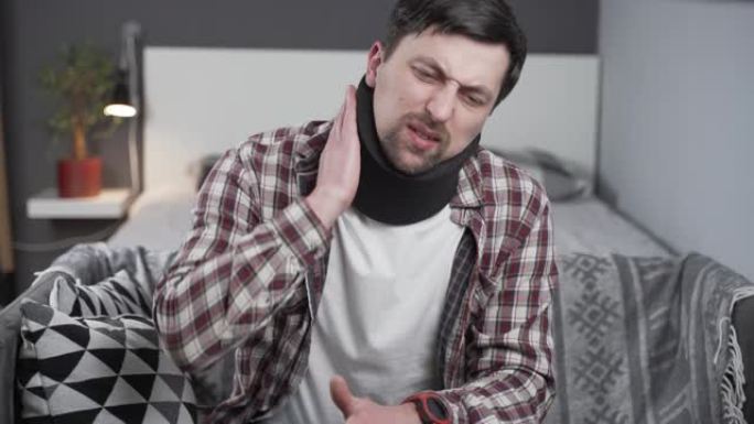 在家沙发上戴着颈托支撑脖子的家伙。颈部受伤的高加索人使用项圈支撑来缓解疼痛。颈椎支架中的男性可减轻脊