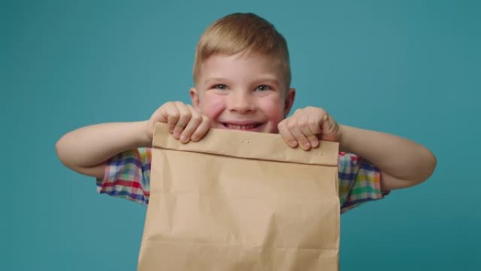 快乐的孩子拿着带食物的纸递送袋，看着站在蓝色背景上的相机。男孩对快餐盒感到兴奋。