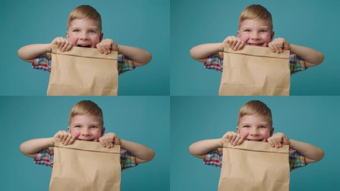 快乐的孩子拿着带食物的纸递送袋，看着站在蓝色背景上的相机。男孩对快餐盒感到兴奋。