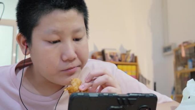 亚洲男生吃炸鸡，在家通过智能手机学习在线课程，新型冠状病毒肺炎，社交疏远的生活方式概念。