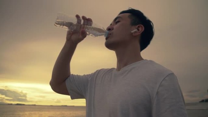 亚洲运动员运动员站在海滩上喝水美丽的日落。运动员训练运动锻炼保健概念。