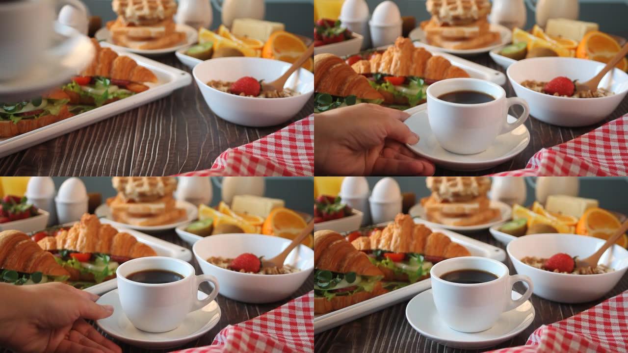 女人的手把一杯咖啡放在桌子上。附近的烤盘配有羊角面包三明治，火腿、奶酪和蔬菜、煮鸡蛋、什锦蔬菜和烤面