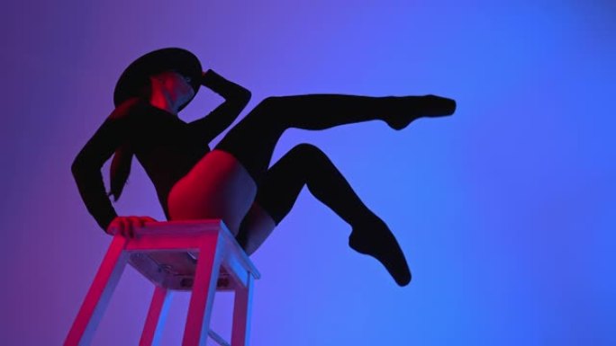 戴着帽子的专业芭蕾舞演员坐在紫色霓虹灯下的多色背景上的高脚椅上。性感性感的芭蕾舞演员穿着长筒袜跳舞。