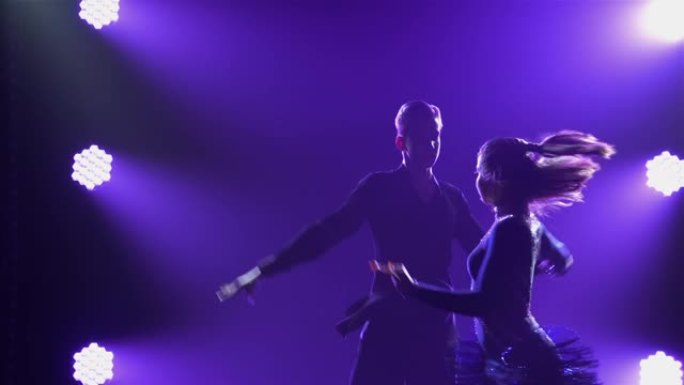 在拉丁美洲舞蹈计划中舞厅舞者的专业表演。在黑暗的烟熏工作室中，紫色灯光背景下的性感夫妇剪影。特写。慢