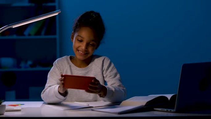 女孩在手机上玩网络游戏，开心地笑。一个女孩在夜灯的灯光下坐在笔记本，教科书和一台打开的笔记本电脑前的