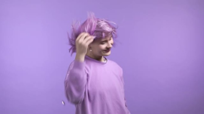 紫色发型的迷人女人在紫色工作室背景上跳舞，她的头发从气流中飘动。穿五颜六色衣服的女孩。积极的情绪。