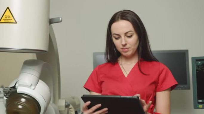身着红色制服的年轻白人女医生的特写肖像，手持数字平板电脑，在泌尿外科医疗中心现代超声波碎石机附近摆姿