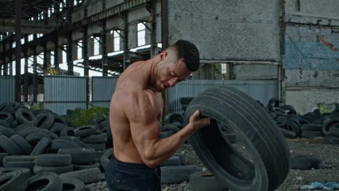 轮胎在废弃处的男运动员。强壮的年轻人在毁坏的建筑背景上进行了激烈的训练，用汽车轮胎训练手臂肌肉。