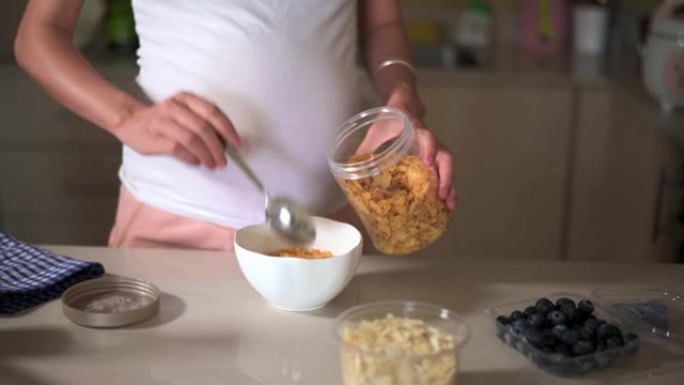 孕妇在家里做早饭玉米片谷类食品，并在厨房里添加配料。制作富含纤维和营养的健康有机餐