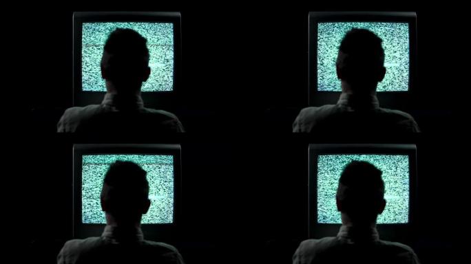 在黑色背景的黑暗演播室里，一个男人坐在闪烁的电视前的轮廓的后视图。老式电视，屏幕上有闪烁的干扰线和一