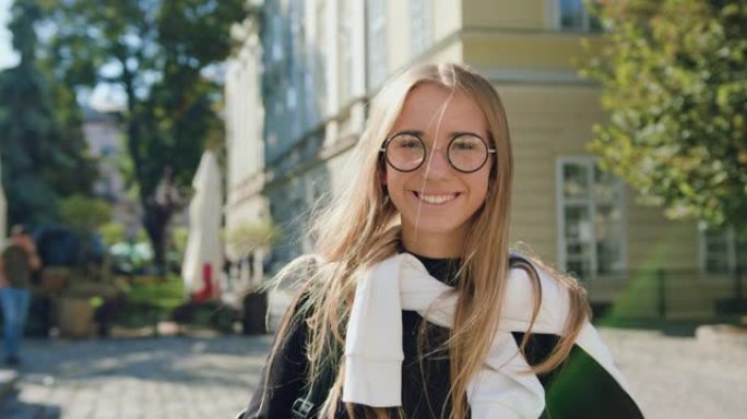 好看的快乐快乐的现代年轻金发女郎戴着眼镜看着相机带着真诚的微笑在阳光明媚的古城建筑的背景下