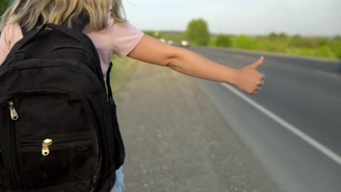一个背着背包的女孩站在路边，竖起大拇指接车。搭便车。用手停车