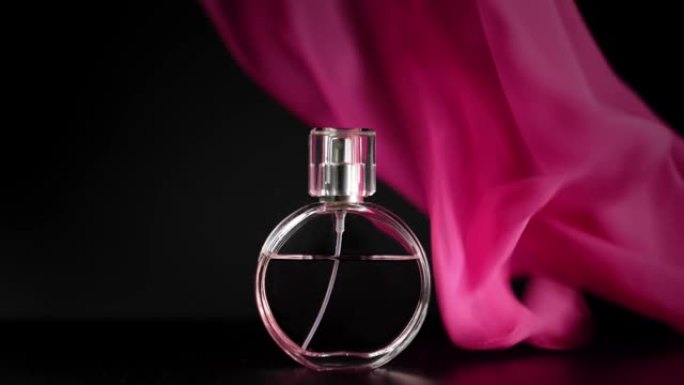 椭圆形的粉红色香水或精油瓶放在桌子上的粉红色布下。粉红色的织物突然从桌子上飘动，暴露出黑色背景下的香