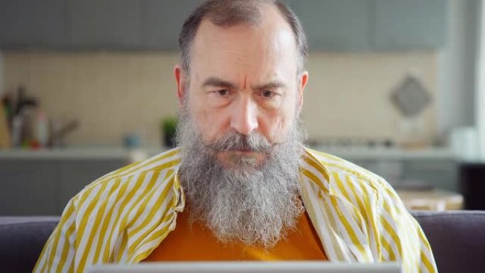 坐在沙发上的笔记本电脑上，有思想的成熟男人的头和肩膀在灰色胡须上打字。男性在家工作，关闭pc并思考