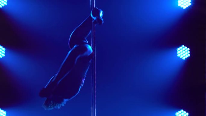 舞蹈演员在一个黑暗的演播室里倒立着绕着钢管旋转。一个细长的运动员身体的剪影在一个黑色的背景与蓝色的光