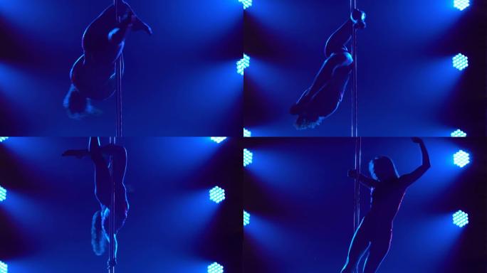 舞蹈演员在一个黑暗的演播室里倒立着绕着钢管旋转。一个细长的运动员身体的剪影在一个黑色的背景与蓝色的光