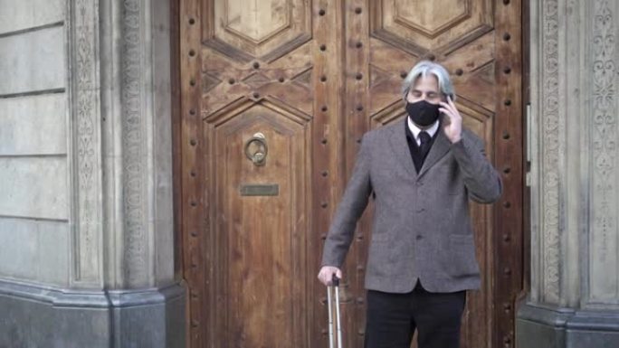 木制酒店大门前戴着口罩和旅行行李的高级商人用手机打电话