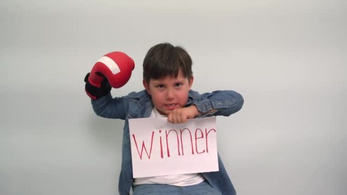 一个戴着拳击手套的男孩拿着一张纸，上面写着题词获胜者严重的孩子在灰蓝色背景上显示了一个带有获胜者一词