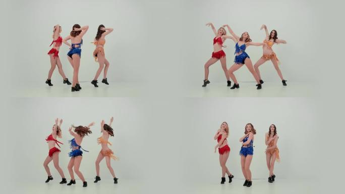三个微笑的女人同步跳舞，移动手臂和臀部，进行垂直分裂。工作室白色背景上闪亮服装的啦啦队团队合作。慢动