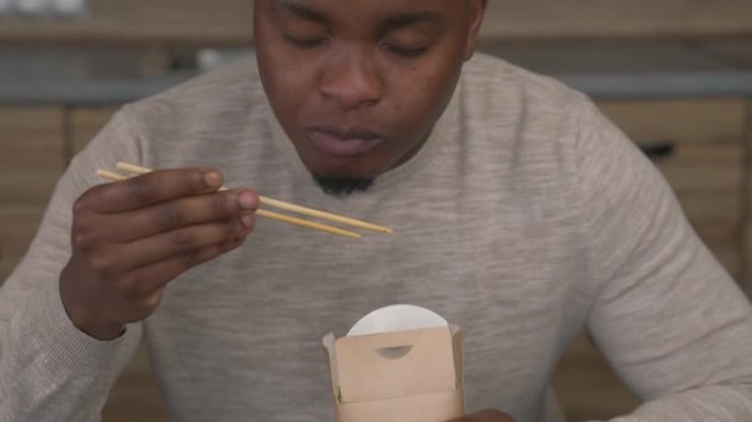 非洲男子坐在餐桌旁，用筷子从盒子里愉快地吃着中国面条。亚洲街头美食。拿走。倾斜摄像机
