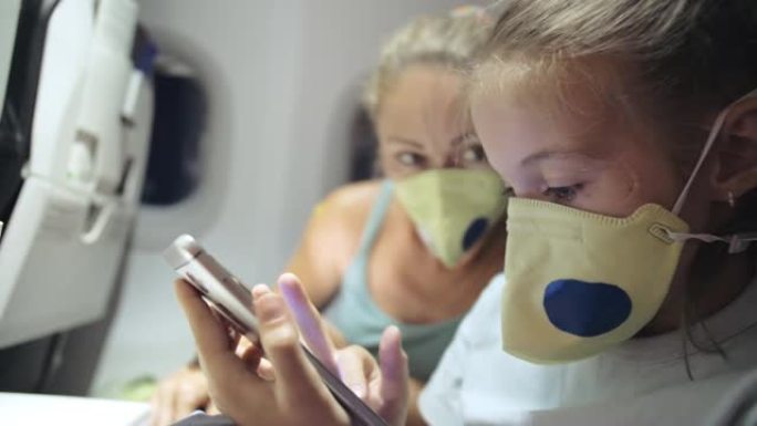 戴着防护医用口罩的飞机上的家庭妇女和儿童婴儿旅行游客高加索人。使用智能手机移动。新型冠状病毒肺炎