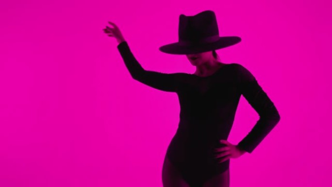 当代舞蹈家戴帽子的女人在彩色灯光下在粉色背景上摆姿势。职业芭蕾舞演员穿着性感的蛇蝎美人服装，独唱表演