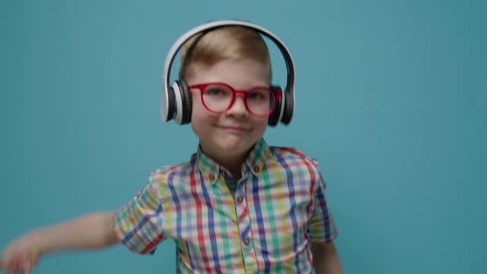 戴着眼镜和耳机的相当可爱的男生在慢动作的蓝色背景上跳舞。快乐的孩子看着相机跳舞。