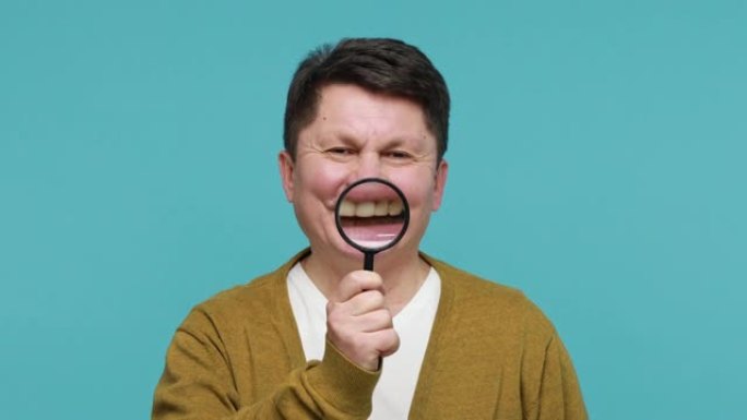 穿着白色t恤和羊毛衫的有趣的中年黑发男子在嘴里拿着放大镜，展示健康的牙齿和口腔医学