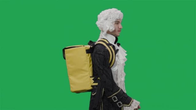 穿着黑色复古西装和假发的朝臣绅士肖像，带着黄色大背包冰箱行走。年轻人在工作室摆姿势，绿屏背景。特写。