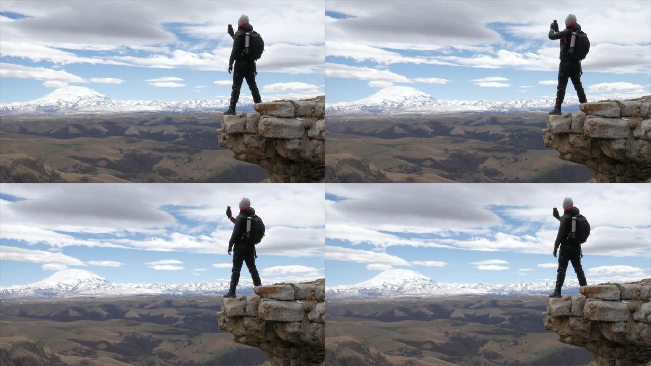 一名男性游客在俯瞰高加索山脉山脉的小山上，埃尔布鲁斯大胆地站在一块岩石上，用手机拍照