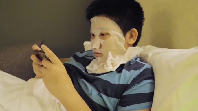 快乐的亚洲男孩用面膜遮住脸，晚上在酒店用智能手机玩网络游戏，生活方式概念。
