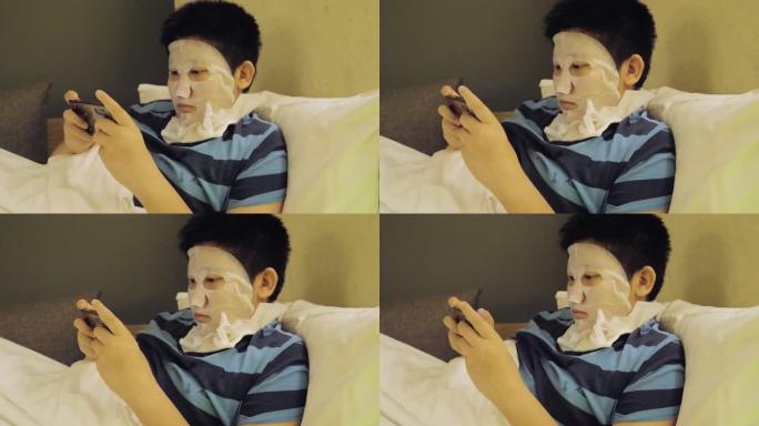 快乐的亚洲男孩用面膜遮住脸，晚上在酒店用智能手机玩网络游戏，生活方式概念。