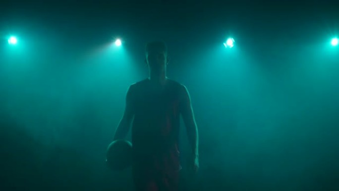 在黑暗和烟雾中，家伙的身影直接走进相机，手里拿着篮子球。篮球运动员在工作室摆姿势，背蓝灯。慢动作准备