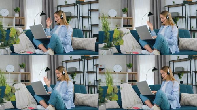 可爱的自信聪明的20岁戴耳机的浅色头发女孩坐在家里的沙发上的莲花姿势，和朋友在网上视频会议，慢动作
