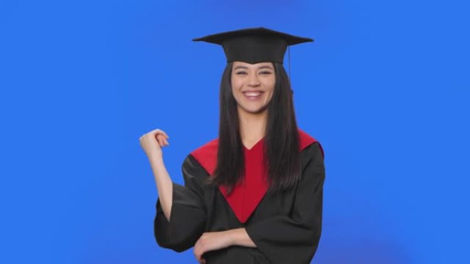 穿着帽子和礼服毕业的女学生的肖像专心地听着，点点头，用手指指着观众。布鲁内特在蓝色屏幕背景上摆姿势。