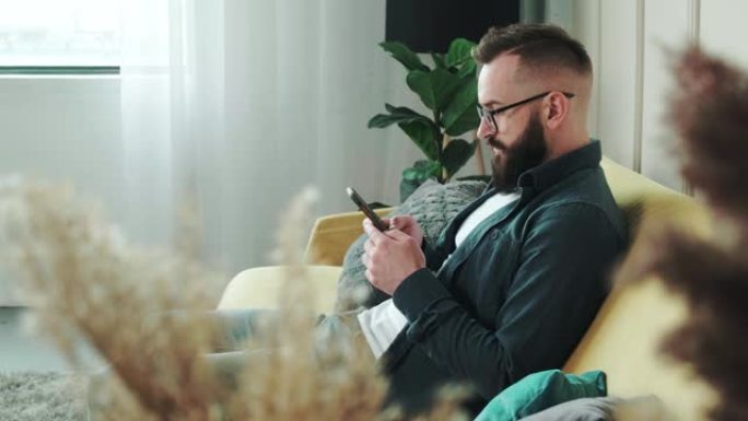 大胡子的男人坐在客厅的沙发上聊天，用手机打字。商人在家通过电话解决业务问题。新一代沉迷于设备概念