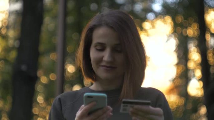 拿着信用卡和电话的学生在街上，黑发女人坐在长凳上，通过智能手机上的银行卡通过互联网进行在线支付
