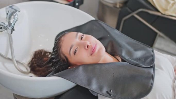 年轻女子躺在沙龙洗发床上，由发型师或美发师在发廊洗头的肖像。美丽的女孩顾客从美容理发店的治疗服务中感