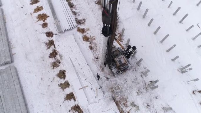 在冬季施工现场工作的桩孔机的俯视图。高冲击重量推动桩深入地面。现场有许多钢筋混凝土桩。冬季阴天