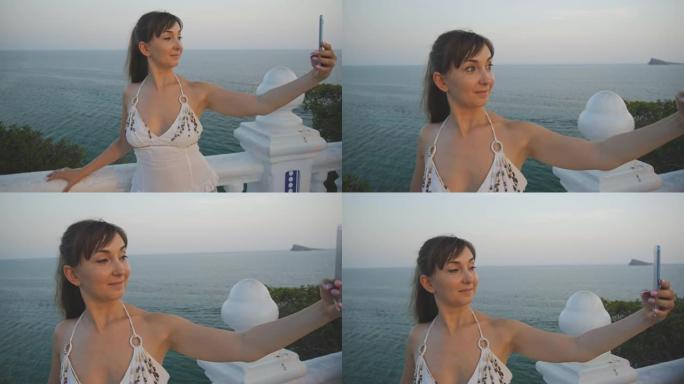 迷人的年轻女子用智能手机自拍。美丽的微笑女孩在夏季日落海边高架阳台上用手机制作自拍照片或视频。