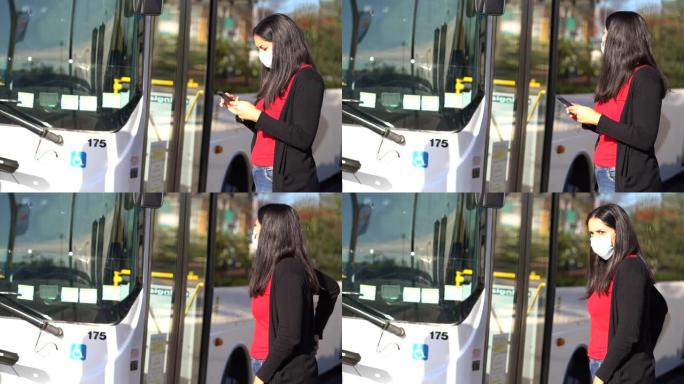 年轻的黑发女子在公共汽车站戴着口罩，看着她的手机。冠状病毒概念
