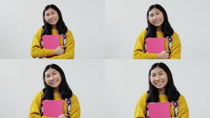 快乐的亚洲女学生穿着黄色毛衣，拿着粉红色的书，看着灰色背景上的相机，生活方式的概念。