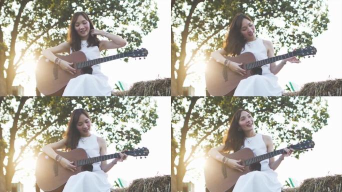 慢动作一位穿着白色连衣裙的年轻年轻亚洲女子在清晨的阳光镜头耀斑下在干草堆上弹吉他。