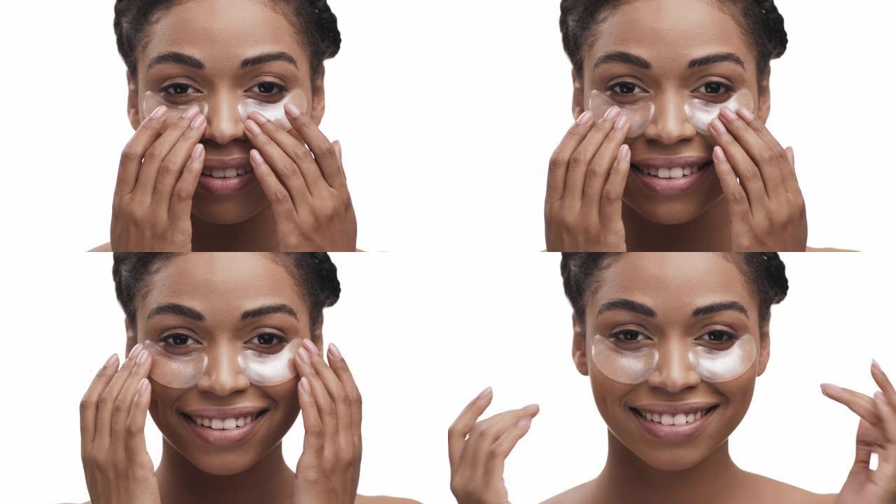 开朗的年轻非洲裔美国女士在眼睛下面应用金色防乏力贴片，对相机微笑，白色背景