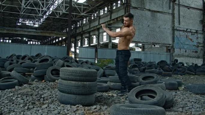 老工厂里拿着重锤的运动员。运动型男子试图用金属锤打破一堆汽车轮胎，对着镜头微笑。在废弃的地方锻炼。