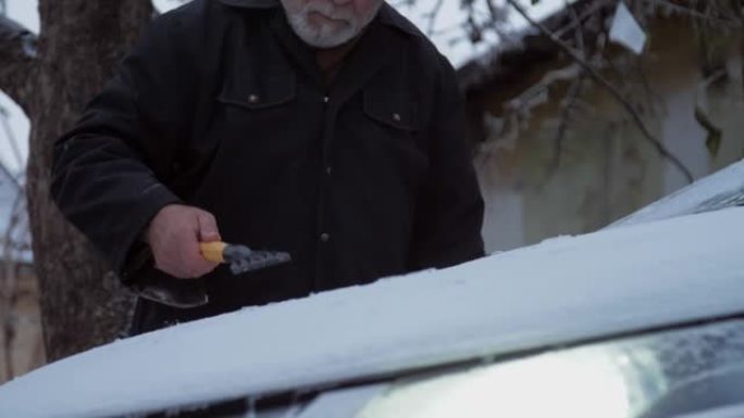 老人用刮冰机从雪地上清洗汽车引擎盖