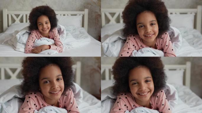 相机放大脸，躺在被子下的非洲裔美国小女孩，看着相机，微笑着。穿着睡衣的少女躺在卧室的白色床上。慢动作